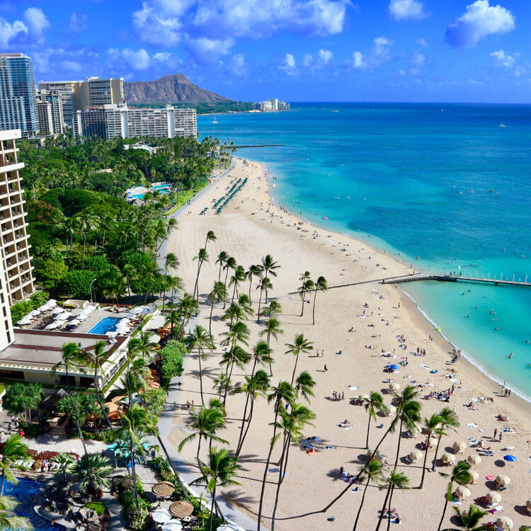 Blick aus der Vogelperspektive auf Waikiki Beach mit türkisfarbenem Wasser, Sandstrand und Palmen, flankiert von Hotels und dem Diamond Head-Krater im Hintergrund