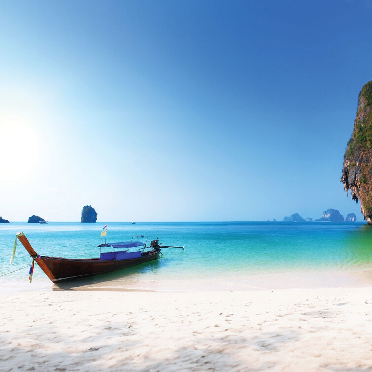 Ein traditionelles Longtail-Boot, das an einem Strand mit kristallklarem, türkisfarbenem Wasser und Kalksteinkarstfelsen festgemacht ist