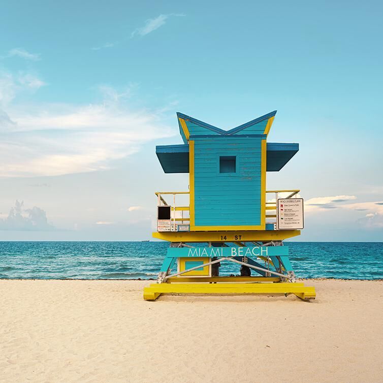 Ansicht von Miami Beach, mit dem Meer im Hintergrund und dem goldenen Sand, mit einem blau-gelben Wachposten und einem Schild mit der Aufschrift „Miami Beach“.