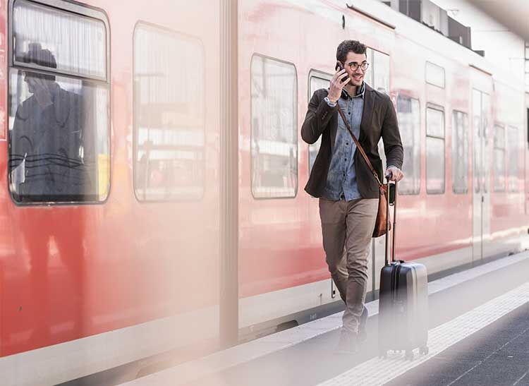 Ein Mann läuft mit seinem Koffer den Bahnsteig entlang, im Hintergrund steht ein roter Zug der Deutschen Bahn