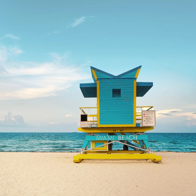 Ansicht vom Strand von Miami Beach, mit dem Meer im Hintergrund und dem goldenen Sand, mit einem blau-gelben Wachtposten und einem Schild, auf dem 'Miami Beach' steht.