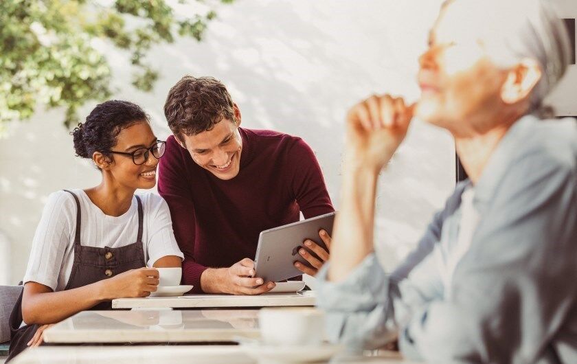 Ein Päarchen sitzt im Café. Sie schauen auf ein Tablet. Eine ältere DAme sitzt rechts weiter vorne allein an einem Tisch. 