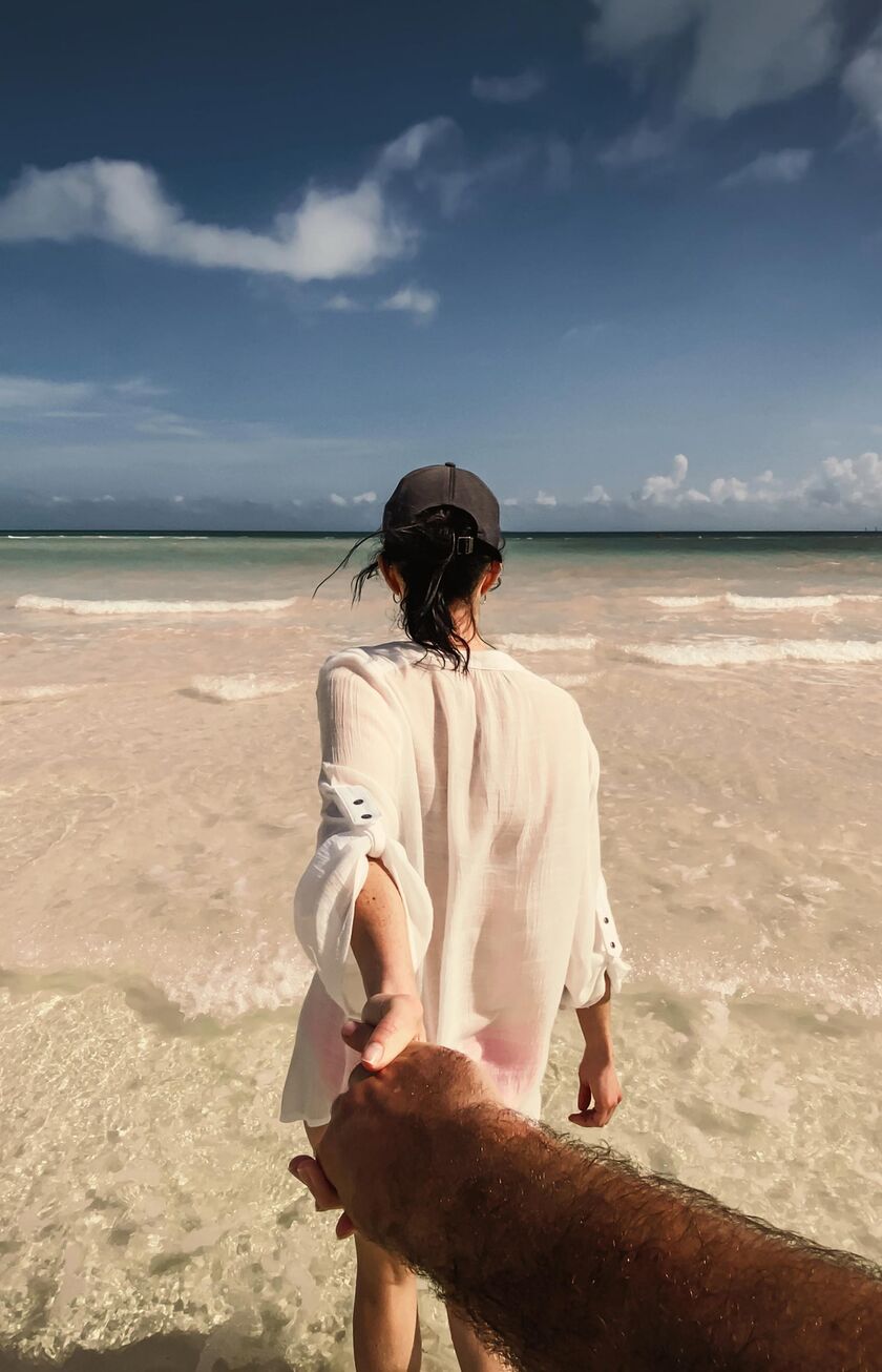 Condor Holidays Frau am Strand von hinten fasst eine Hand, die ins Bild ragt.