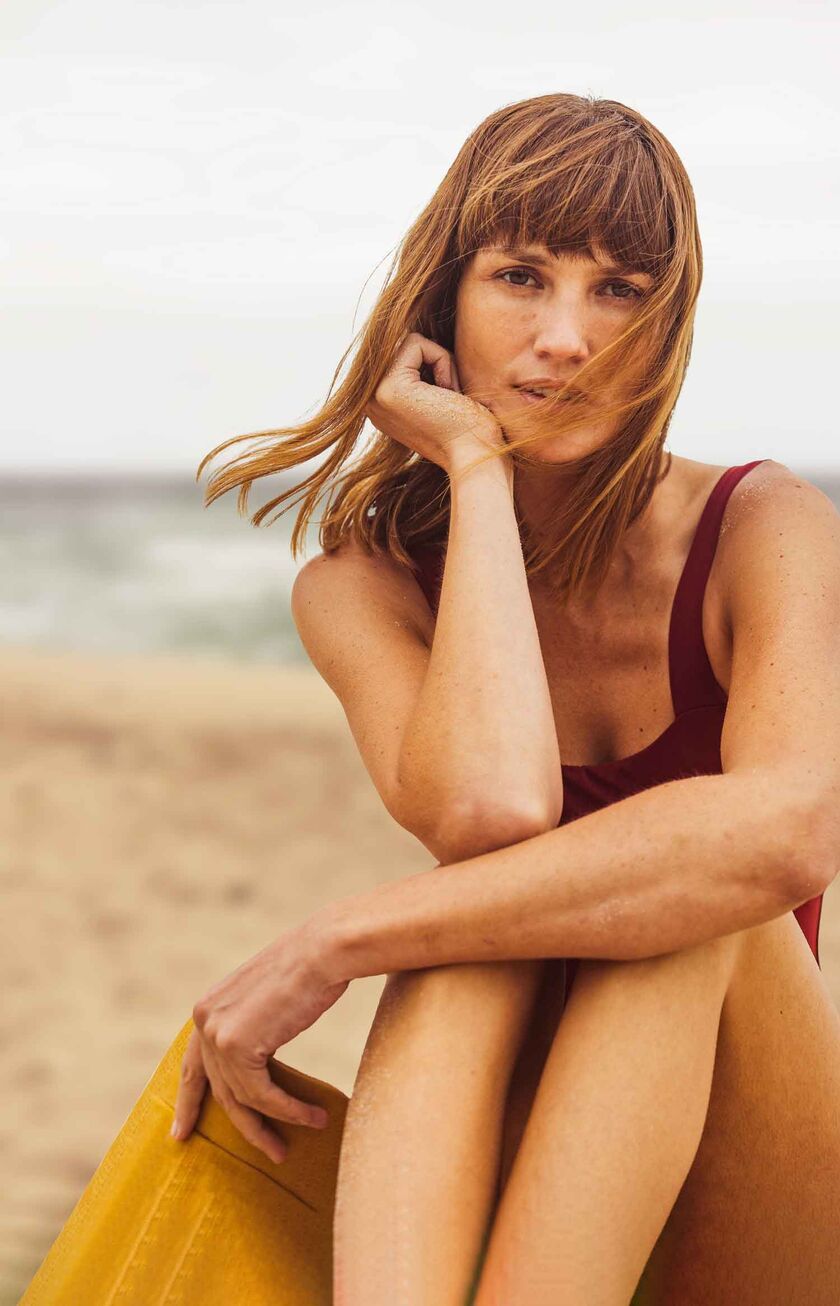 Eine Frau sitzt im Badeanzug am Strand und schaut verträumt in die Kamera