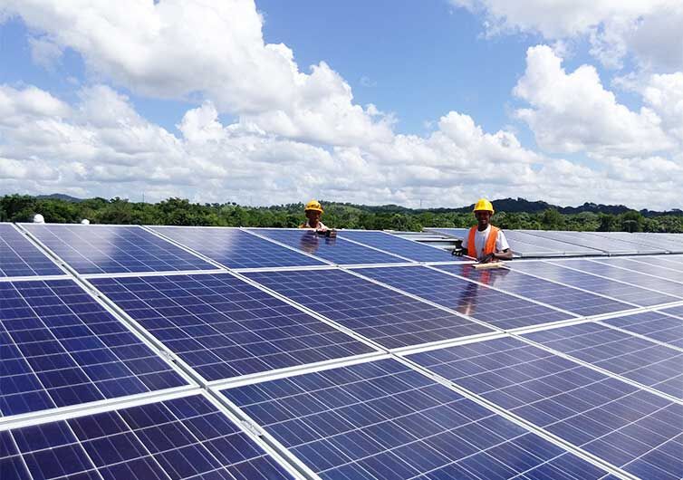 Two workers in safety vests and helmets installing solar panels under a partly cloudy sky in the Dominican Republic.