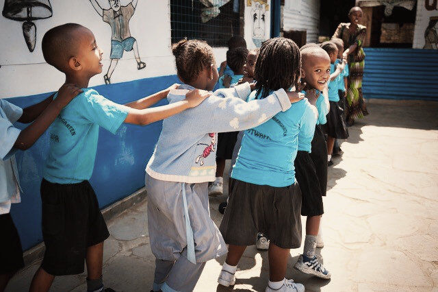 Children holding each other by the shoulders and walking in a line. 