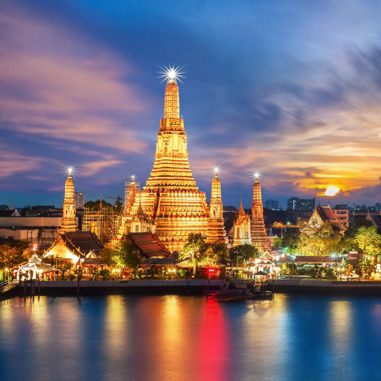 Wat Arun, the Temple of Dawn, stands majestically against a soft pink sky at dusk with the Chao Phraya River in the foreground and birds flying overhead in Bangkok, Thailand.