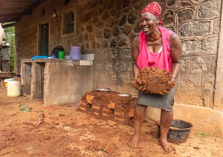 A woman in Kenya smiling while holding a large amount of mud, standing outside her home.