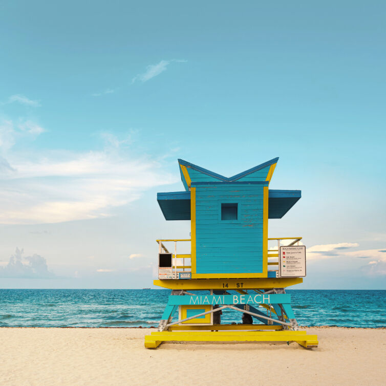 Vue de la plage de Miami Beach, avec la mer en arrière-plan et le sable doré, avec un poste de surveillance bleu et jaune et un panneau où est écrit Miami Beach.