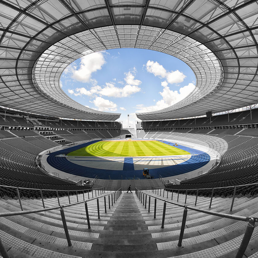 Interior view of the Olympic Stadium in Berlin with a circular open roof, showcasing a sunny sky above and a central emblem or design in the middle.